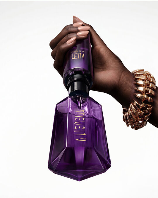Une main tenant le flacon emblematique violet de l'eau de parfum Alien, sur un fond blanc uni.