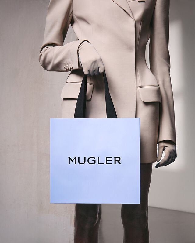 Une personne vetue d'un costume beige et de gants, tenant un sac a provisions bleu clair muni de poignees noires, debout devant un mur gris clair texture.