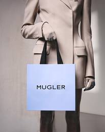 Personne vetue d'un costume beige et de gants, tenant un sac a provisions bleu clair avec des poignees noires, debout devant un mur gris clair texture.
