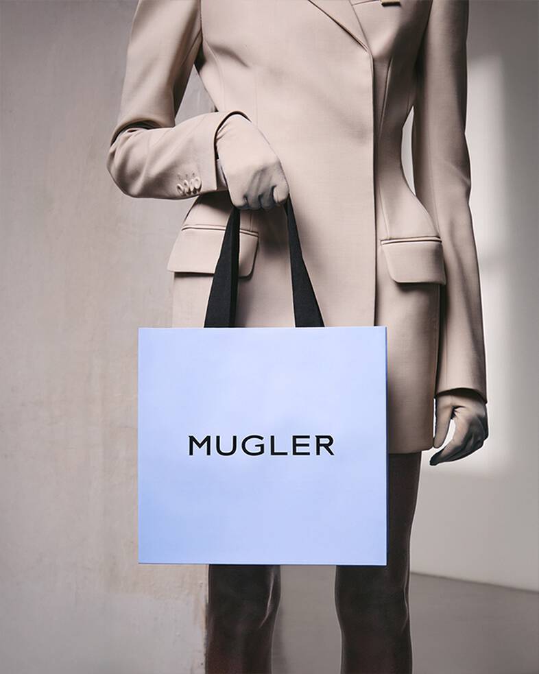 Une personne vetue d'un costume beige et de gants, tenant un sac a provisions bleu clair muni de poignees noires, debout devant un mur gris clair texture.
