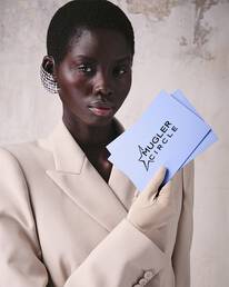 Une personne a la peau foncee et aux cheveux courts, vetue d'un blazer couleur creme et d'une boucle d'oreille en maille argentee, tenant deux cartes bleu clair dans une main gantee.