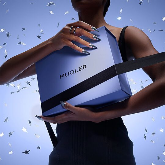 Une personne tenant une boite bleue de marque Mugler, entouree de confettis en forme d'etoiles, avec des ongles longs et stylises.