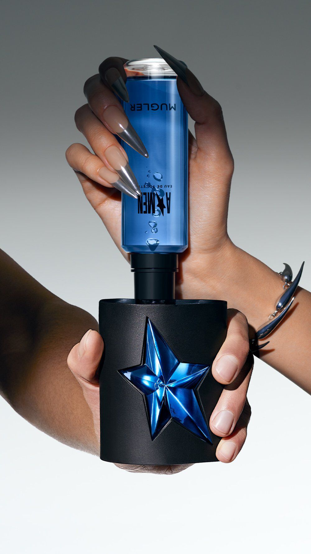 Une main tient un flacon de parfum, avec un bouchon bleu en etoile, sur fond degrade gris clair.