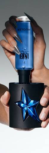 Une main tient un flacon de parfum, avec un bouchon bleu en etoile, sur fond degrade gris clair.