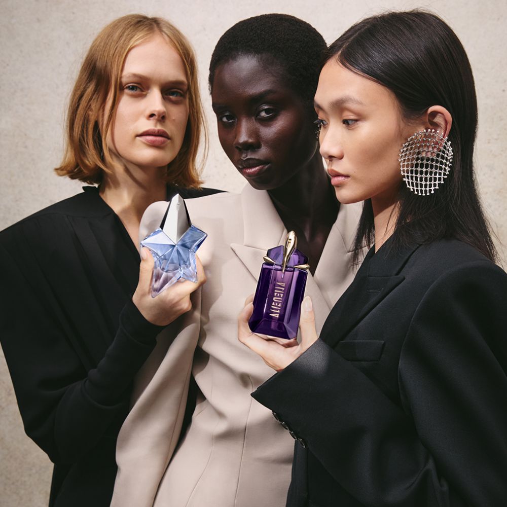 Trois femmes tiennent des flacons de parfum bleu et violet. Le fond est un mur beige