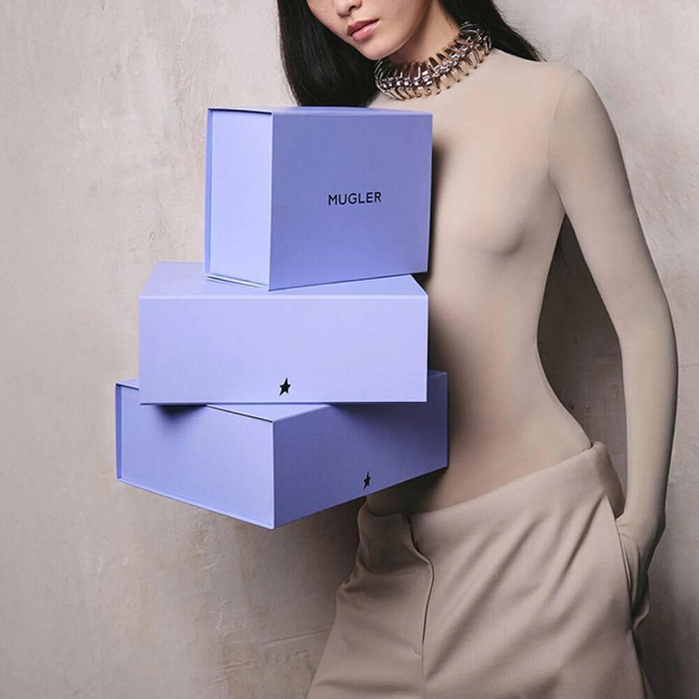 Une femme tenant une pile de trois boites-cadeaux Mugler bleu violet ornees du logo en forme d'etoile.