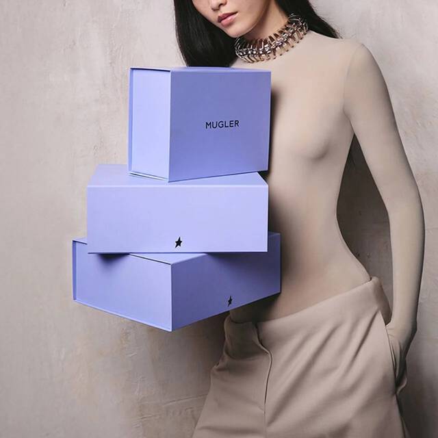 Une femme tenant une pile de trois boites-cadeaux Mugler bleu violet ornees du logo en forme d'etoile.