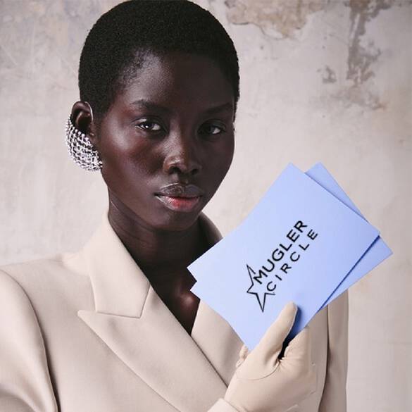 Une personne a la peau foncee et aux cheveux courts, vetue d'un blazer couleur creme et d'une boucle d'oreille en maille argentee, tenant deux cartes bleu clair dans une main gantee.