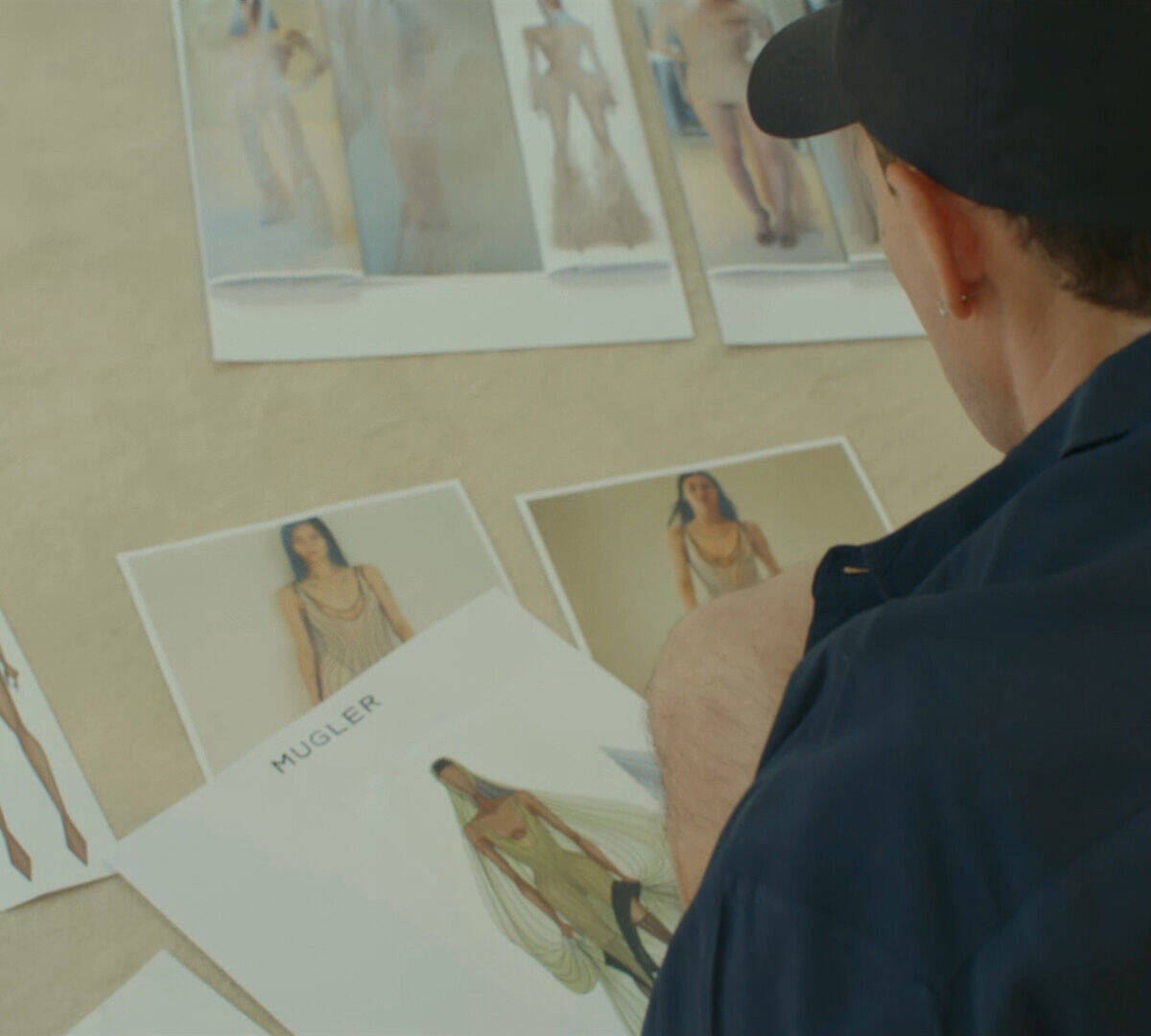 Une personne vetue d'une chemise bleu fonce et d'une casquette noire examine plusieurs croquis de mode et des photographies de mannequins sur une surface claire.