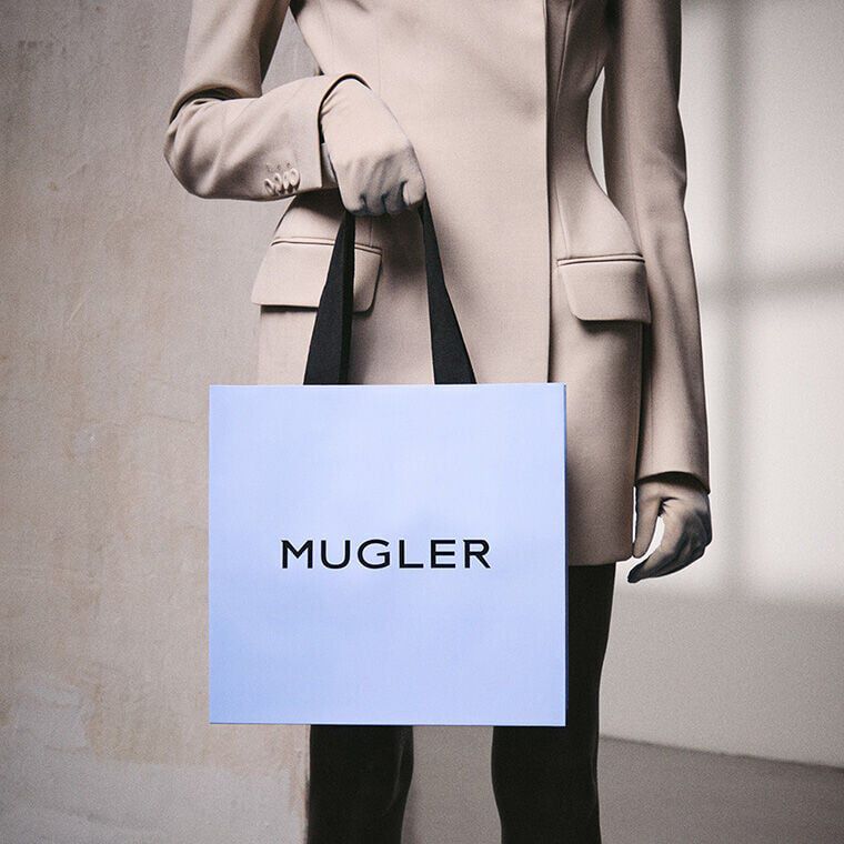 Une personne vetue d'un blazer Mugler sur mesure et de gants, tenant un sac de courses Mugler bleu clair a anses noires, symbole d'un cadeau de luxe.