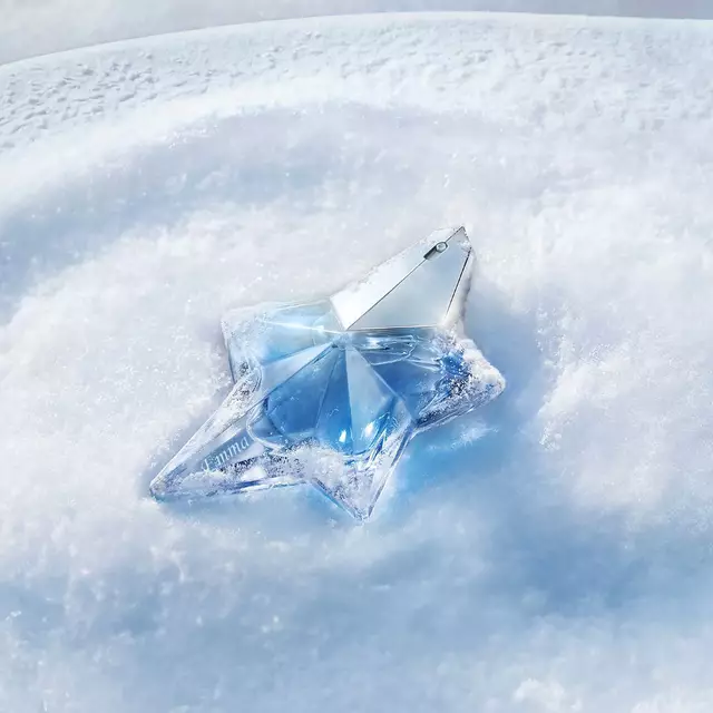 Gros plan sur un flacon de parfum Mugler Angel emblematique, de couleur bleu clair et en forme d'etoile, reposant avec elegance sur un lit de neige d'un blanc immacule et etincelant, sous une douce lumiere bleue. Le texte indique : « Faites graver votre cadeau. Personnalisez-le grace a notre service de gravure gratuit. »