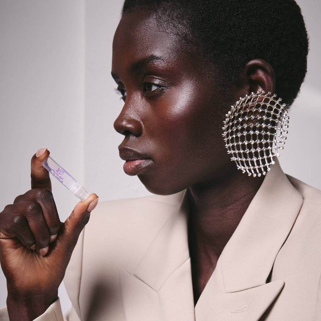 une femme a al peau noire habille en beige qui tient un echantillon de parfum