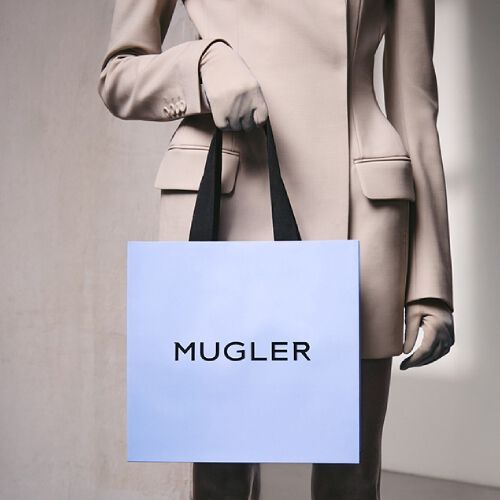 Personne vetue d'un costume beige et de gants, tenant un sac a provisions bleu clair avec des poignees noires, debout devant un mur gris clair texture.