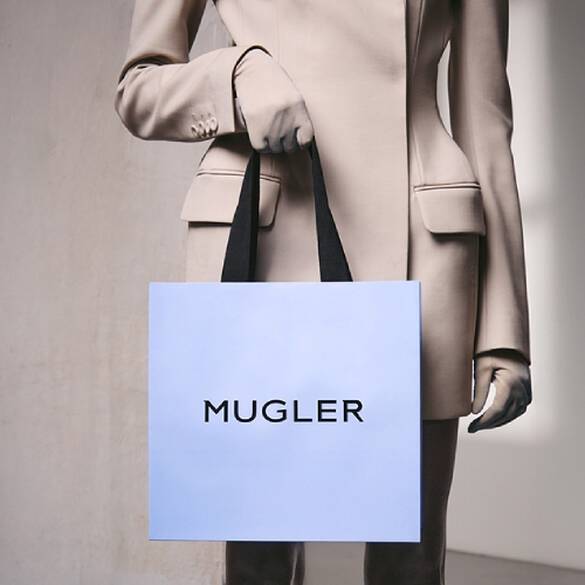 Personne vetue d'un costume beige et de gants, tenant un sac a provisions bleu clair avec des poignees noires, debout devant un mur gris clair texture.