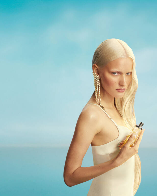 Nouvelle banniere publicitaire pour la campagne « Starlicious les eaux gourmandes » de Mugler, mettant en scene un mannequin aux longs cheveux blonds et aux yeux bleus, vetu d'une robe jaune pale, tenant le flacon du parfum « Pineapple Musk » devant un fond de ciel bleu clair. Ces parfums sont delicieux, a collectionner et a superposer.