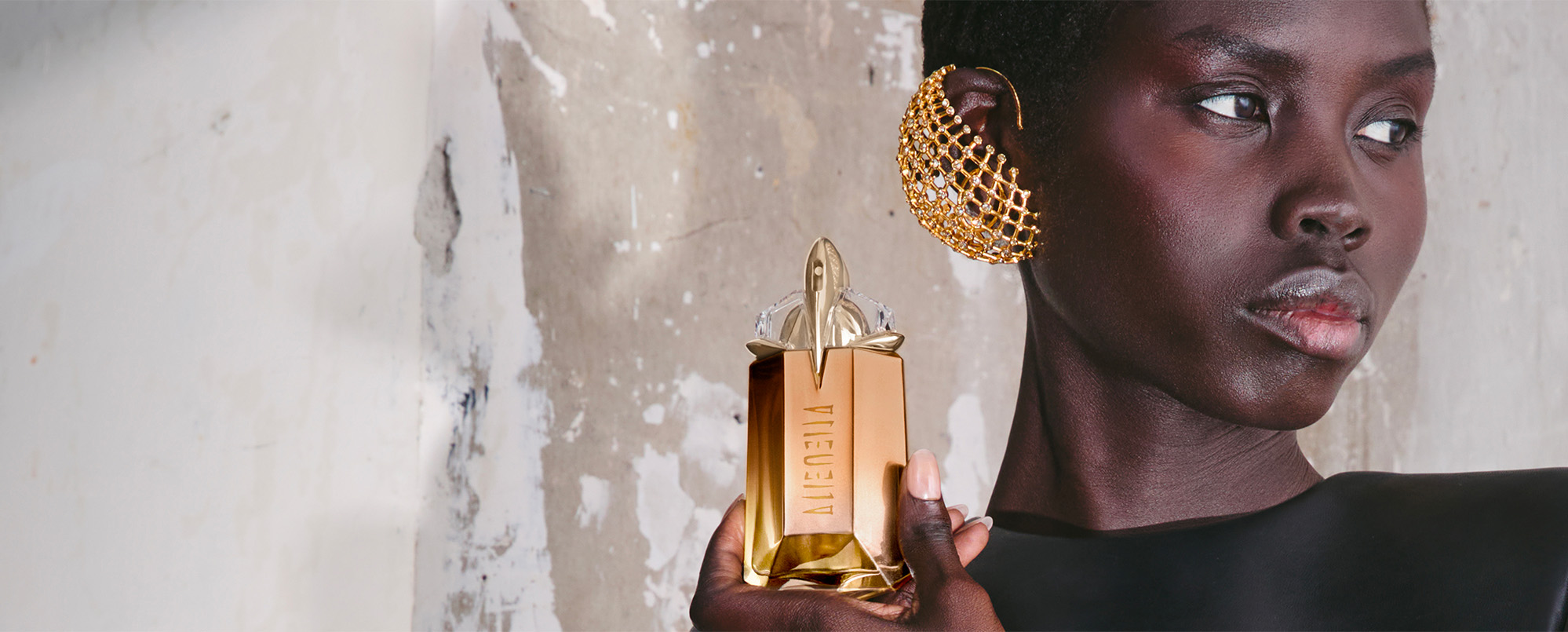 Une élégante mannequin en noir tenant le flacon talisman couleur pêche et or de Mugler Alien Goddess Supra Florale , photo lifestyle pour la campagne publicitaire d'un parfum de luxe.