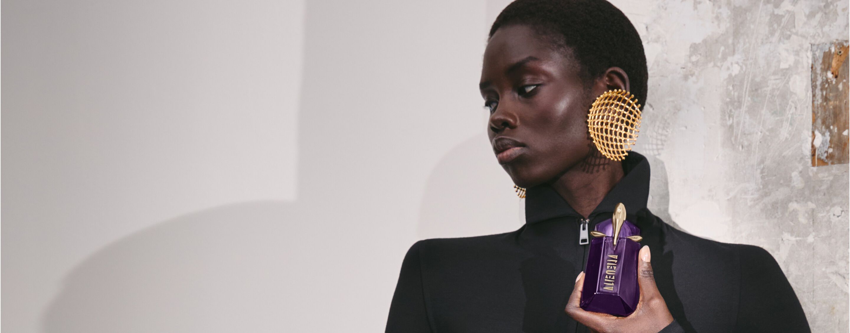 une femme habillée en noir avec la tête de profil tenant un flacon de parfum violet dans sa main devant un mur gris