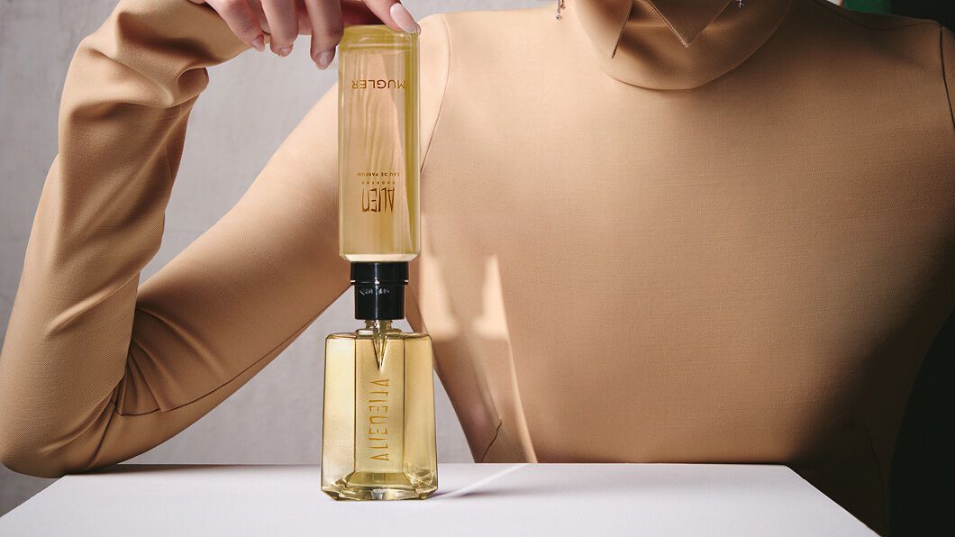 un flacon de parfum doré remplit par une recharge jaune tenu par la main d'une femme habillé en beige