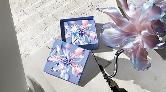 Bannière de la campagne Mugler pour la fête des mères 2026, mettant en scène les flacons emblématiques des parfums Angel et Alien dans un paysage floral céleste et parisien, rendant hommage aux mères extraordinaires.