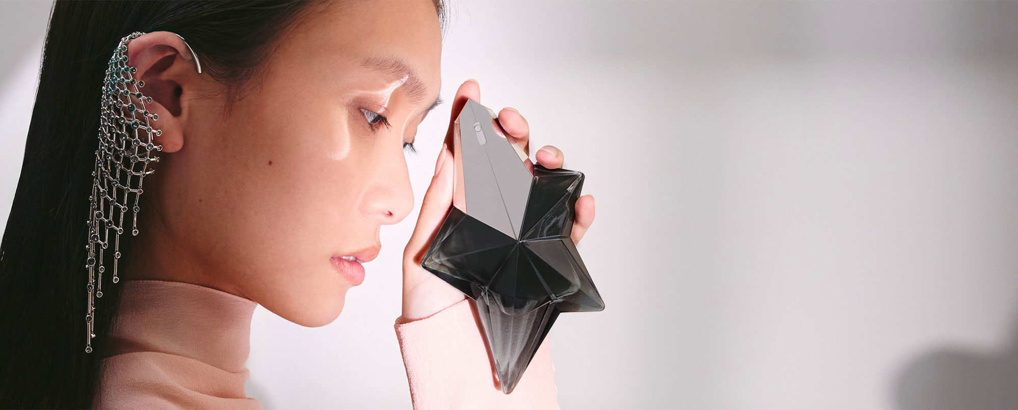 Photo illustrant un style de vie luxueux et sensuel : une femme appliquant le parfum Mugler Angel Fantasm, présenté dans un flacon en forme d'étoile, derrière son oreille.