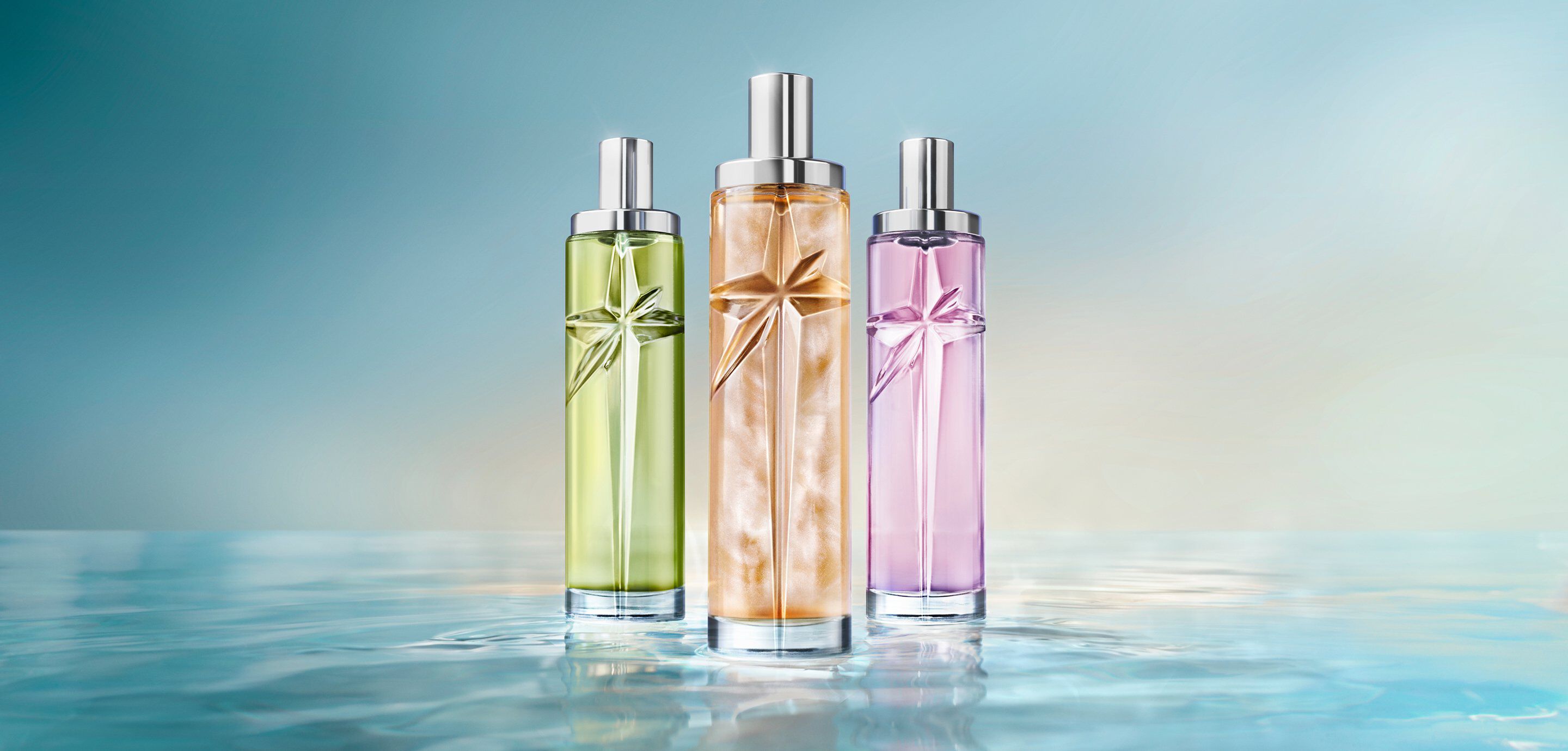 La nouvelle collection Starlicious : trois grands flacons de parfum cylindriques aux bouchons argentés, déclinés en vert, ambre doré et violet lavande, alignés côte à côte sur une surface réfléchissante évoquant l'eau, sur un fond bleu clair.