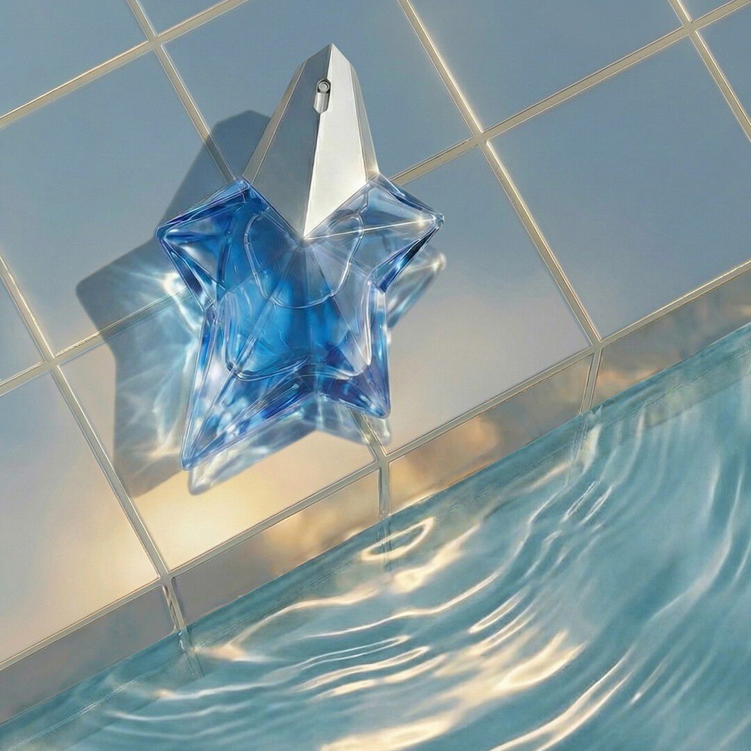 Le flacon en forme d étoile de l'eau de parfum Mugler Angel, de couleur bleue, repose au bord d un bassin aux eaux ondulantes, sur un fond de carreaux pâles et brillants.