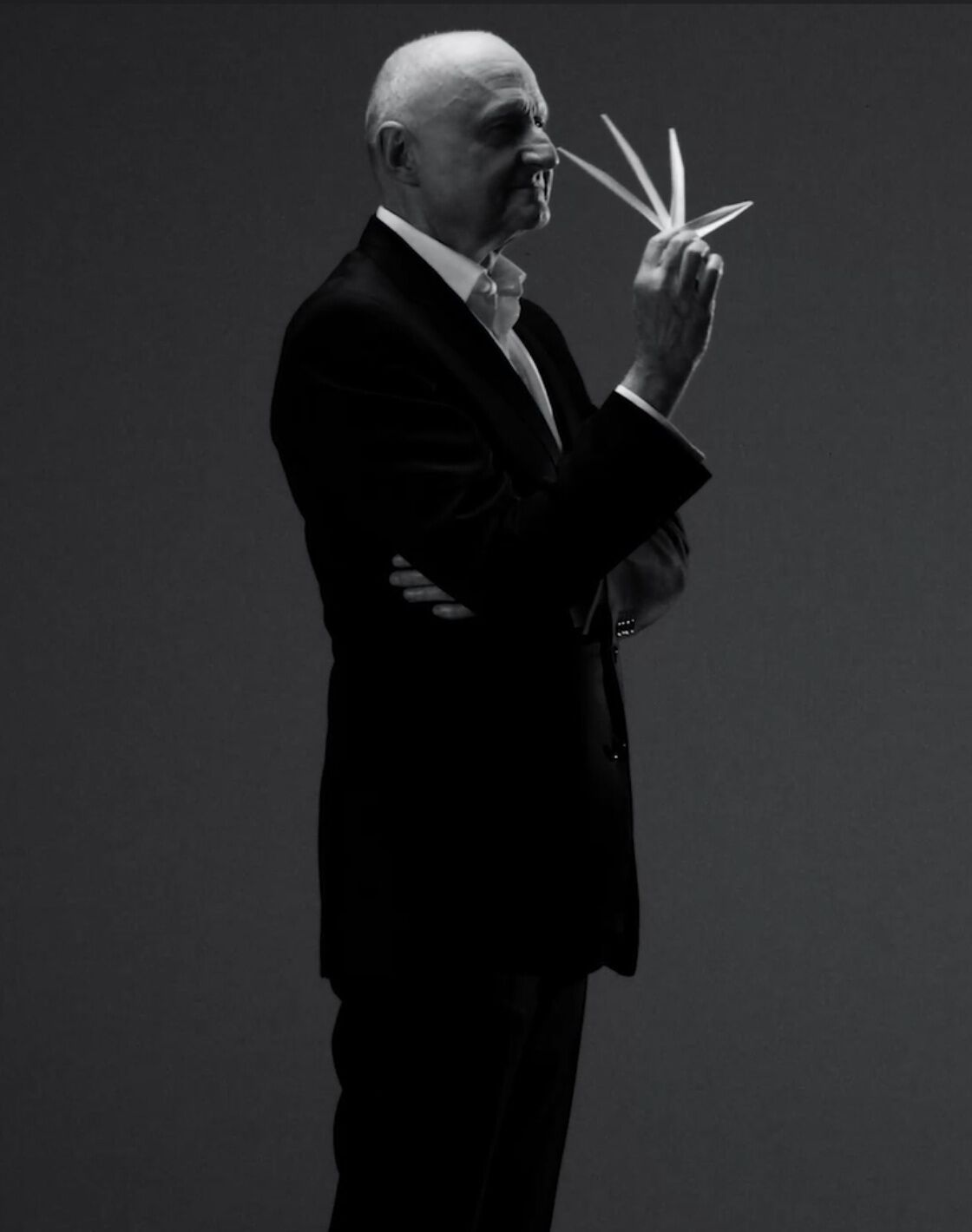 Homme en costume noir tenant des feuilles, posé dans une lumière monochrome, avec une expression réfléchie.