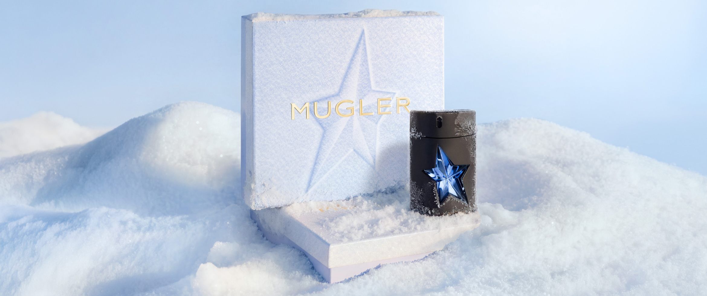 Un coffret cadeau de Noël Mugler, de couleur bleu glacier et à la texture originale, trône dans la neige à côté d un flacon noir épuré d eau de toilette A*Men orné d une étoile bleue découpée.