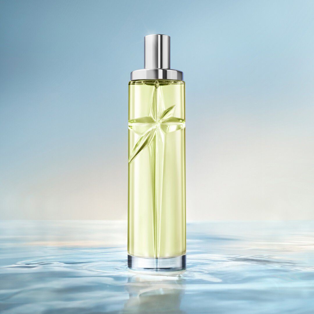 Un flacon de recharge vert Mugler Starlicious posé dans une eau peu profonde et miroitante, sur un fond bleu lumineux.