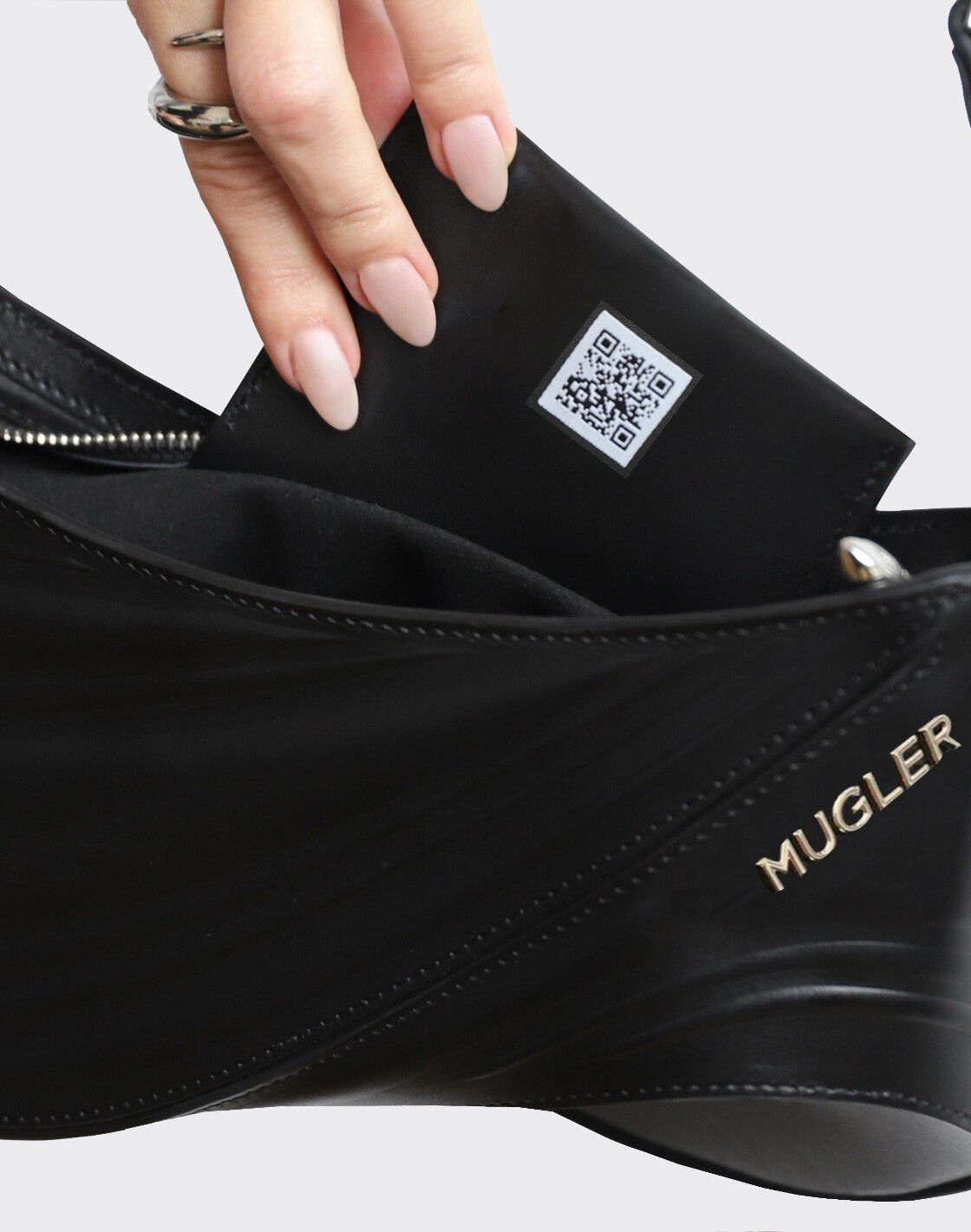 Main dans un sac noir Mugler, sortant un petit objet avec un code QR. Ongles manucurés en nude.