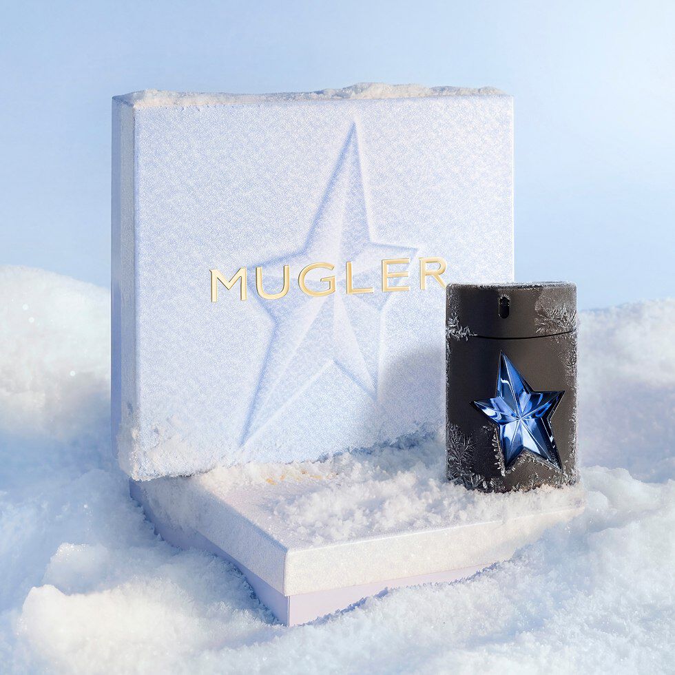 Un coffret cadeau de Noël Mugler, de couleur bleu glacier et à la texture originale, trône dans la neige à côté d un flacon noir épuré d'eau de toilette A*Men, orné d une étoile bleue découpée.