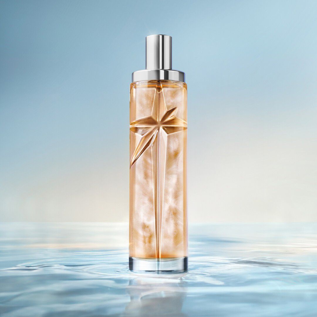 Flacon de recharge Mugler Starlicious Amber posé dans une eau peu profonde et miroitante, sur un fond bleu lumineux.