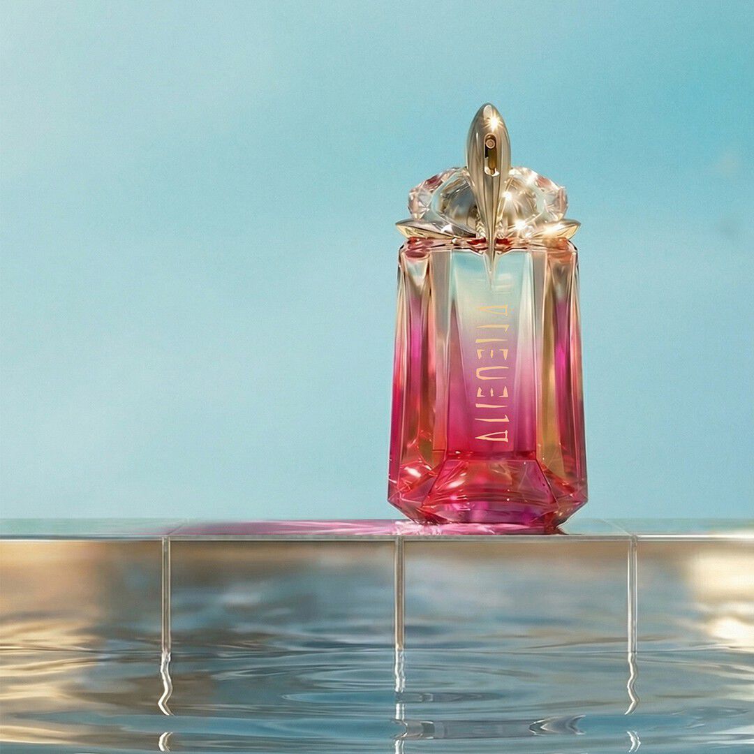 Flacon Alien Pulp de Mugler en verre dégradé rose, posé sur le rebord carrelé d une piscine au-dessus d une eau miroitante, sur fond bleu pâle.