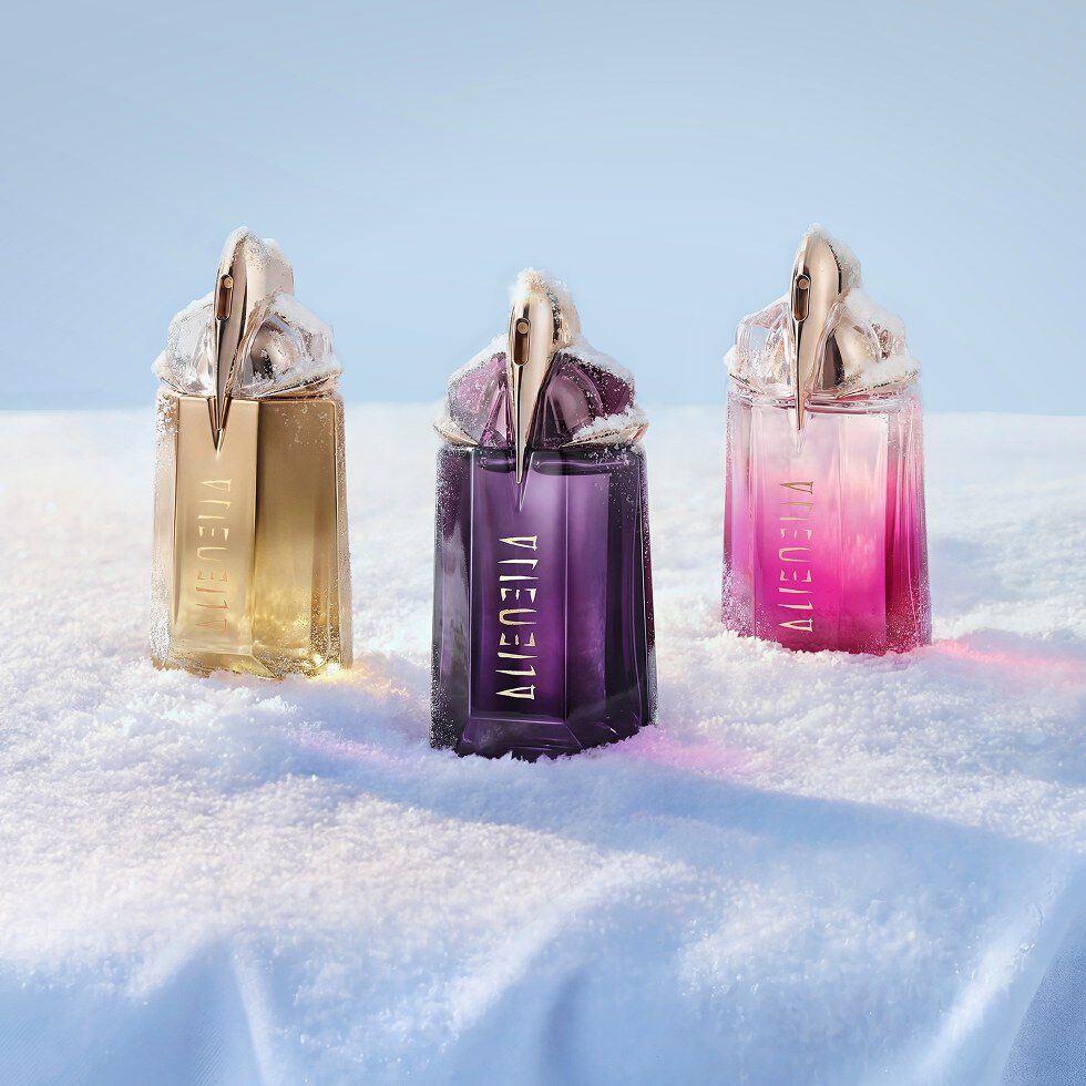 Un trio de flacons du parfum Mugler Alien, l un doré (Alien Goddess), l autre violet foncé (Alien eau de parfum) et le dernier rose vif (Alien pulp), alignés sur un buffet recouvert de neige.
