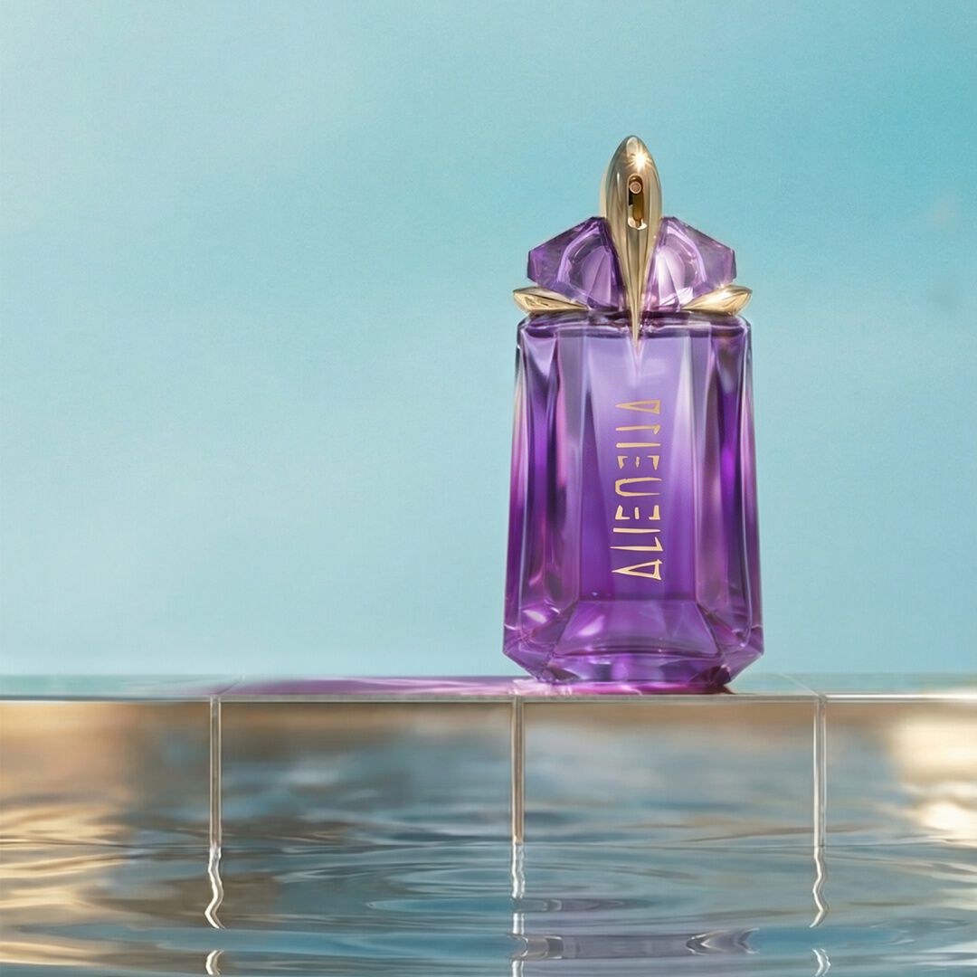 Flacon du parfum Alien de Mugler en verre violet rehaussé de touches dorées, posé sur un socle en verre transparent au-dessus d'une eau miroitante et ondulante, sur fond bleu clair.